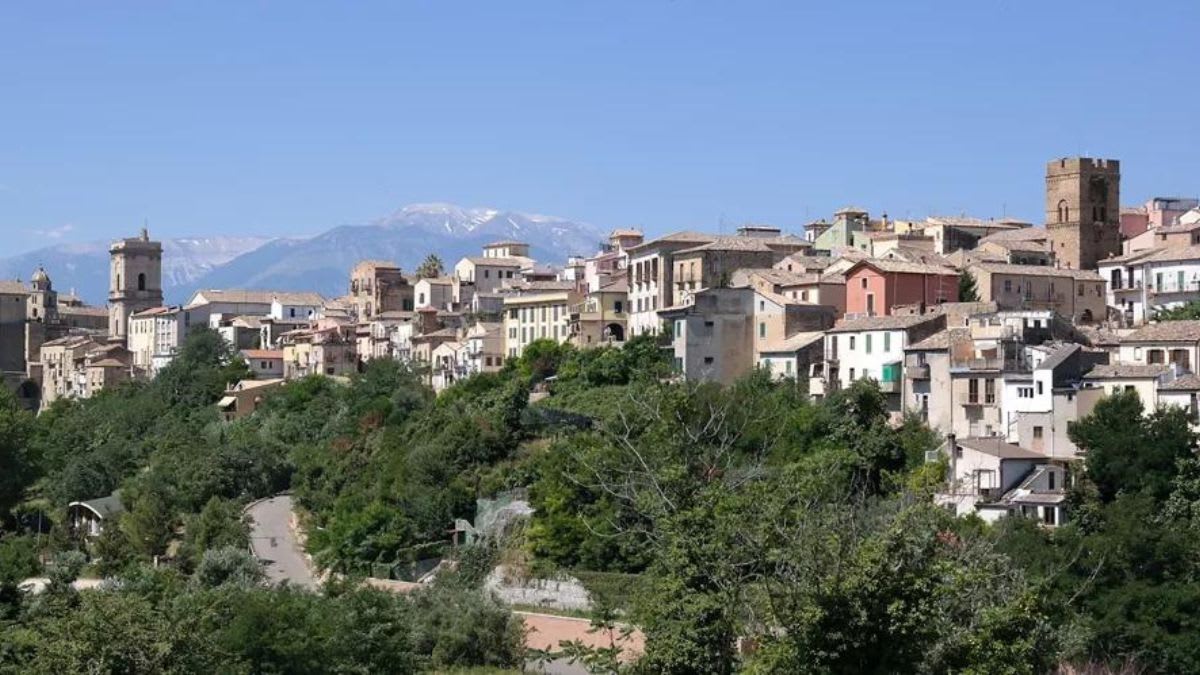 Lush green trees in the foreground frame a picturesque Italian hilltop town with snow-capped mountains in the distant background.
