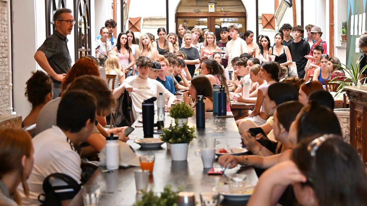 A group of young people are gathered around a long table in the foreground, with a man standing and addressing them in the background.