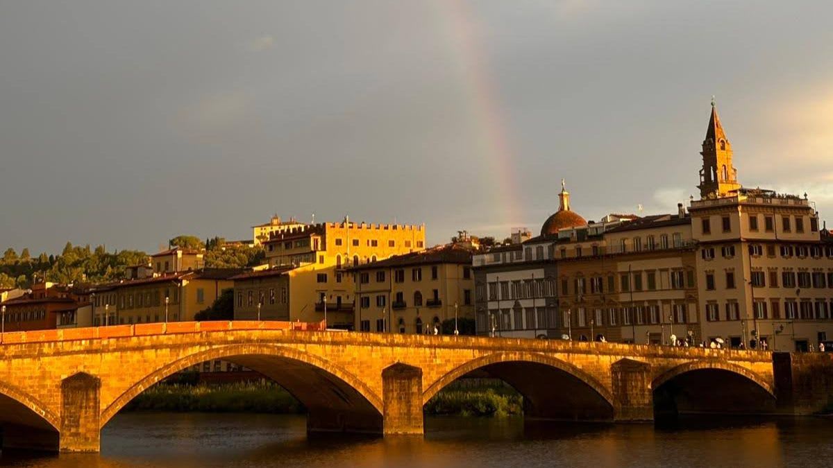 A stone bridge arches over a river in the foreground, with a city skyline and a faint rainbow in the background.