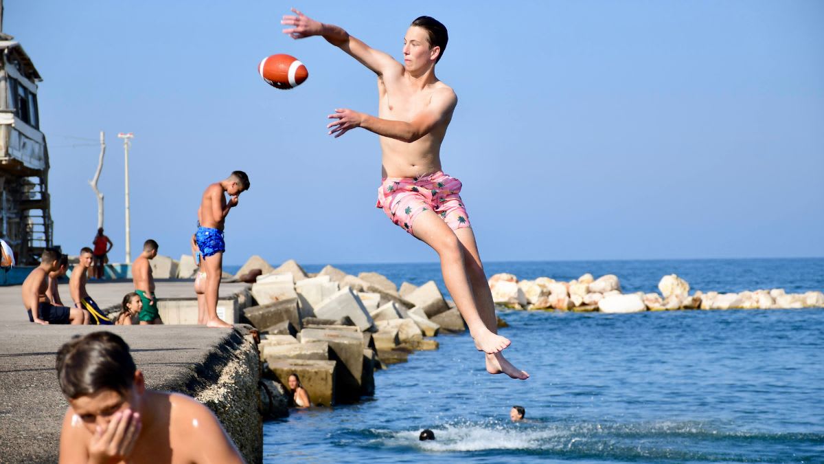 A young man in pink shorts leaps into the blue ocean with a football, while others relax on a concrete pier under a clear sky.