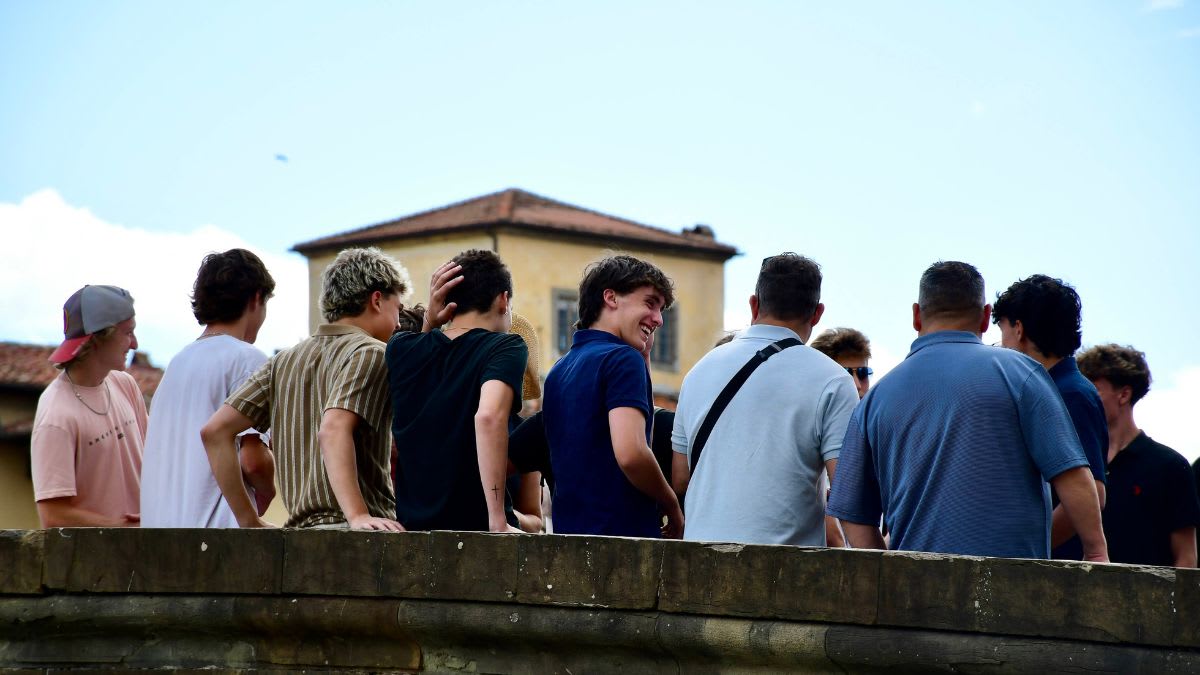 A group of young men are sitting on a stone wall in the foreground, with a building and a cloudy sky in the background.