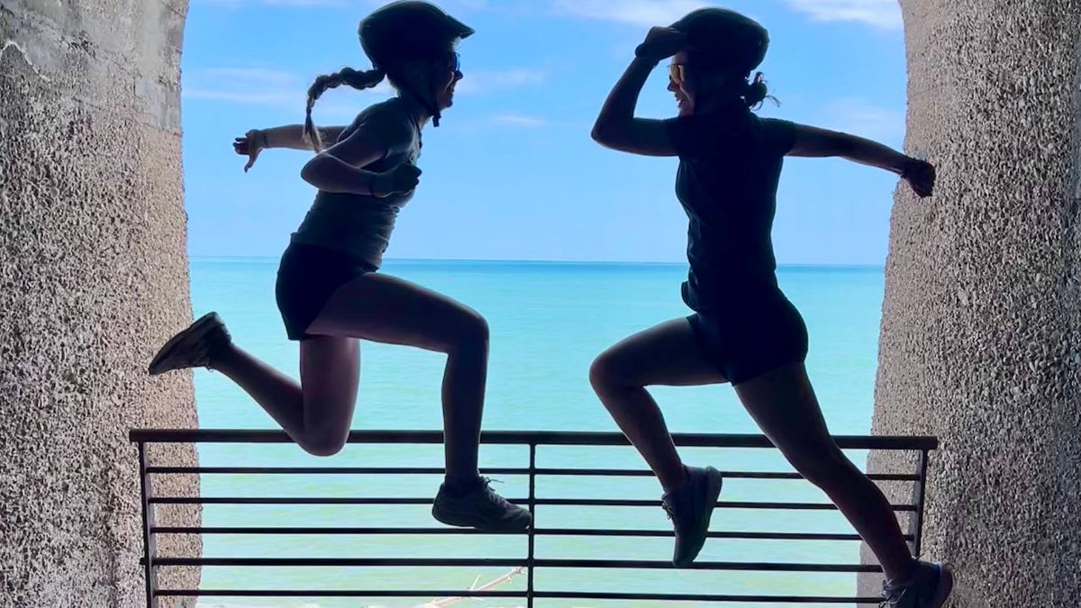 Two silhouetted figures in athletic wear leap joyfully against a bright blue sky and ocean, framed by rough stone archways.