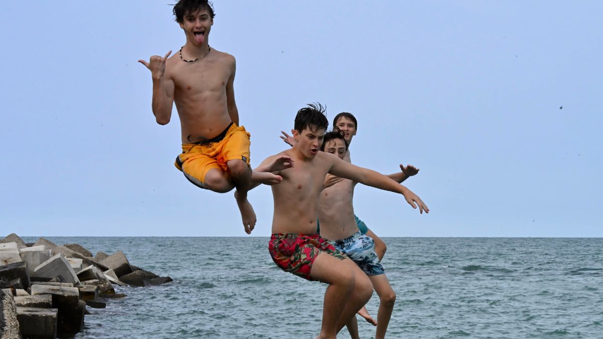 Four boys in swim trunks jump off a rocky breakwater into the ocean under a cloudy sky.