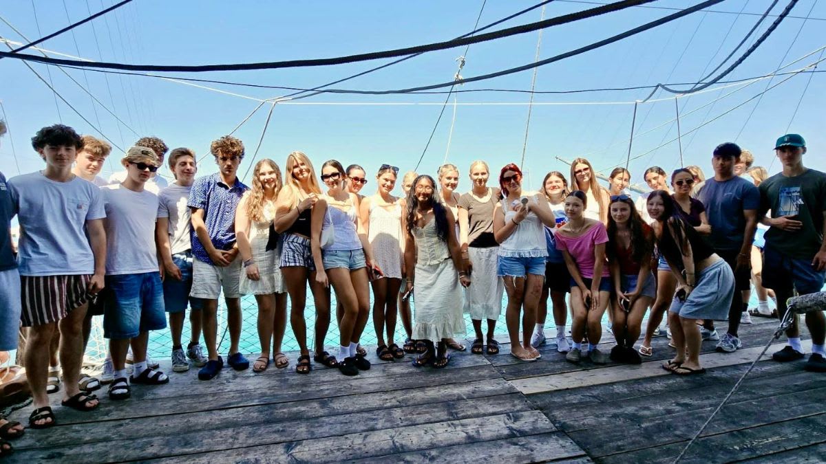 A group of young people stands on a wooden deck with a clear blue sky and calm water in the background.
