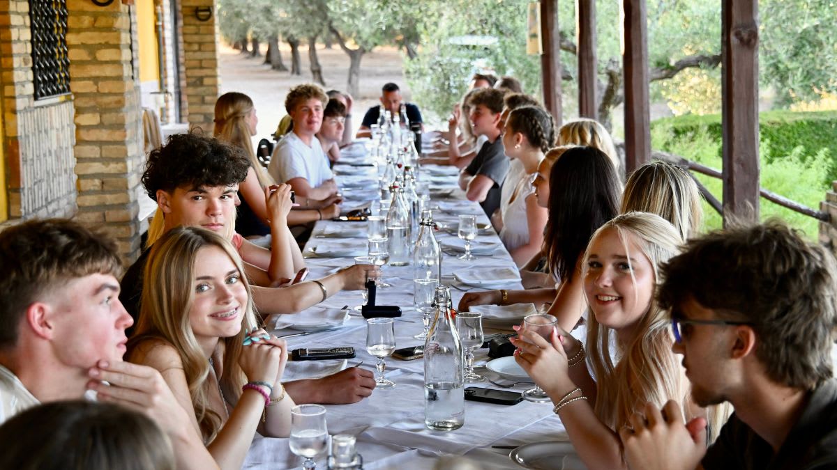 A group of young people are seated at a long, set table in the foreground, with a tree-lined path and greenery in the background.