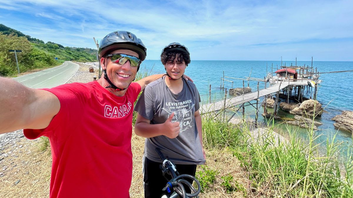 Two smiling people in helmets pose for a selfie in the foreground with a stilt house on rocks in the ocean and a coastal road in the background.