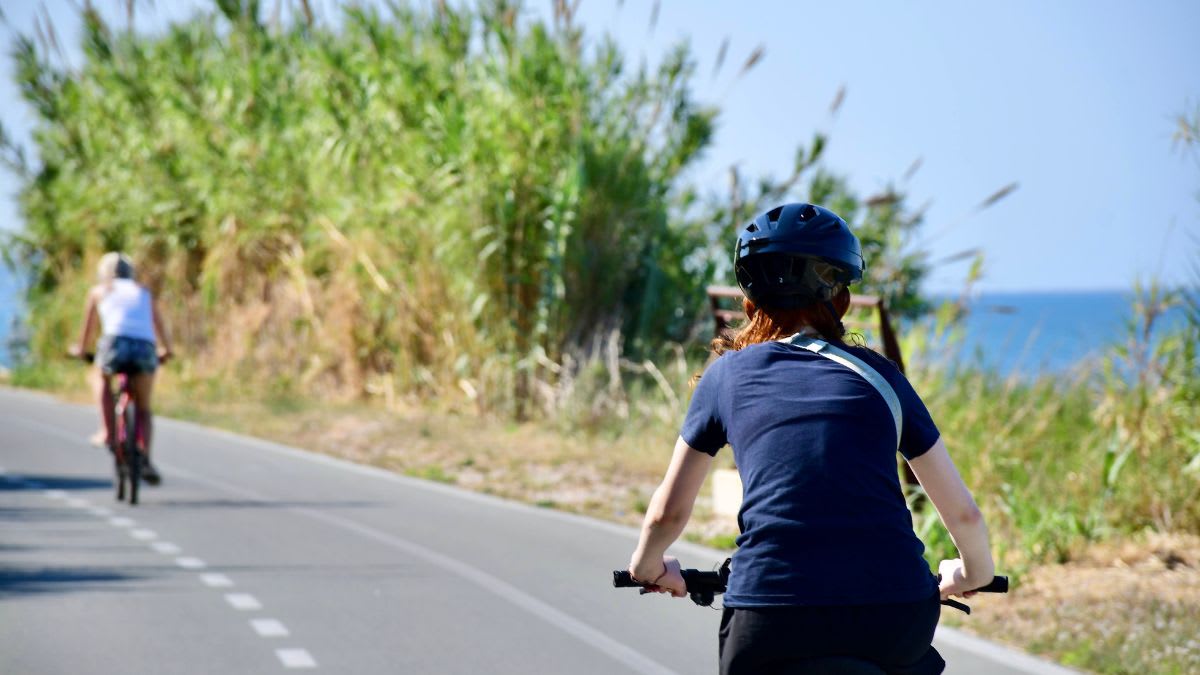 Two cyclists ride on a paved path with lush green foliage to their left and the blue ocean in the distance.