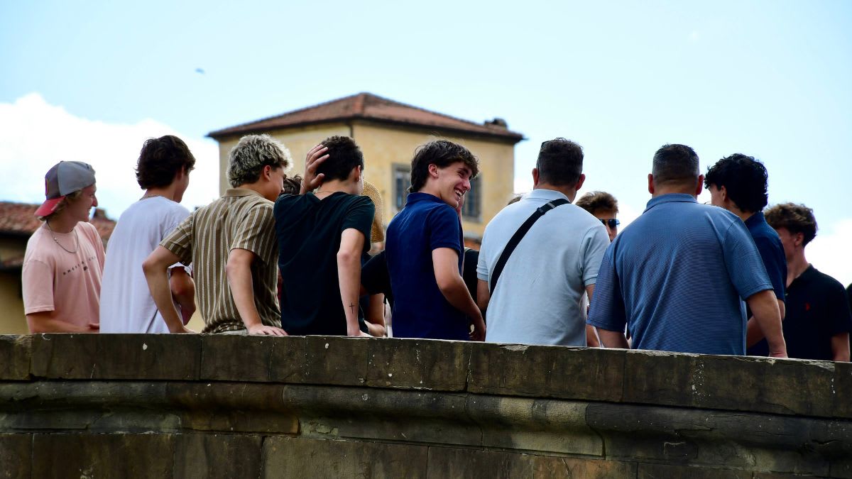 A group of young men stand on a stone bridge in the foreground, with a building and sky visible in the background.