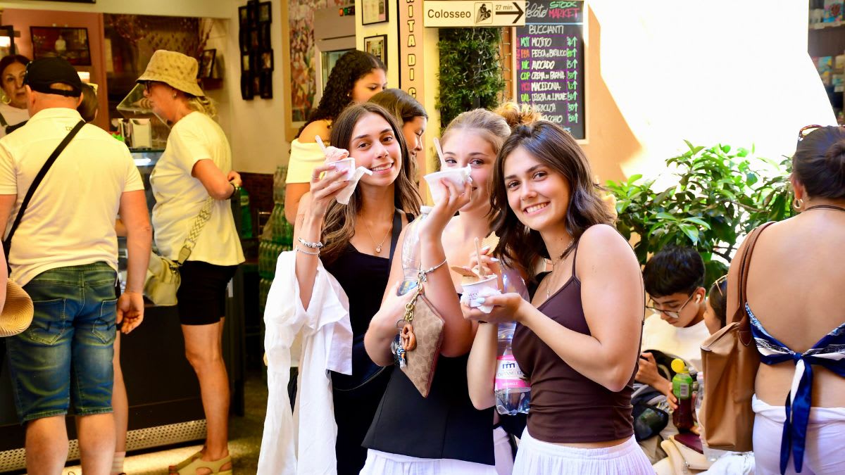 Three young women in the foreground are enjoying ice cream on a sunny day, while in the background, a crowd of people are gathered outside a shop with a sign pointing towards the Colosseum.