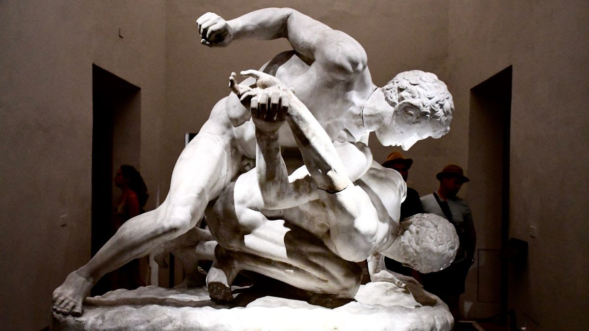 A marble sculpture of two men wrestling is displayed in a dimly lit museum hall with blurred figures in the background.