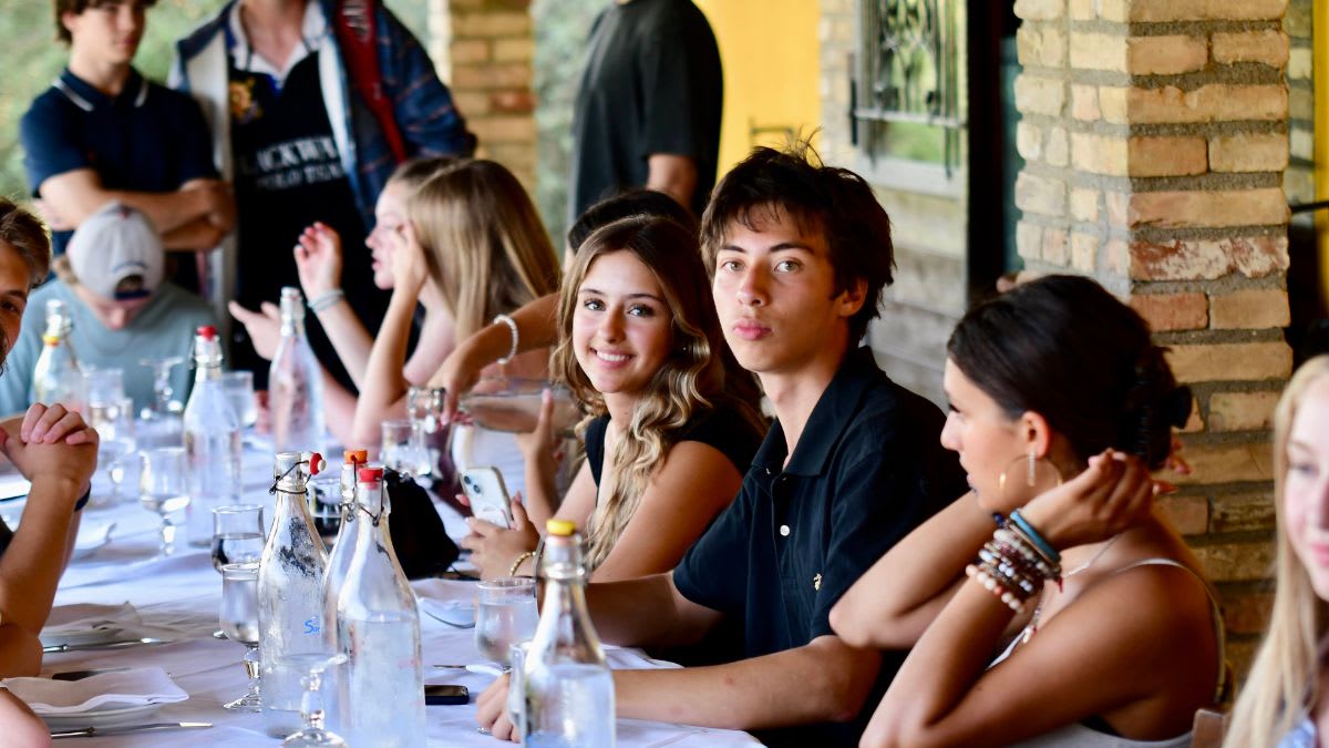 A group of young people are seated at a long table set with bottles and glasses, with a blurred background of people and a building.