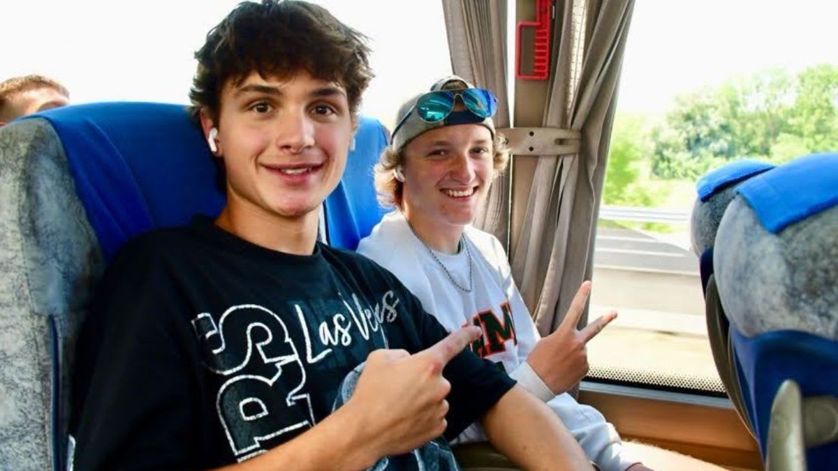 Two smiling young men, one in the foreground pointing at the camera and the other in the background giving a peace sign, are seated on a bus with blurred scenery visible through the windows.