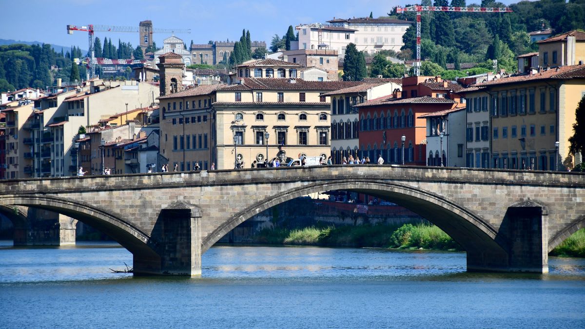 A stone bridge with arched spans crosses a blue river in the foreground, while a cityscape with historic buildings and a distant tower rises in the background under a clear sky.