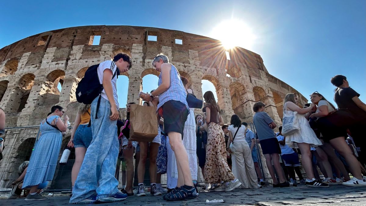 Tourists gather in the foreground with the sun shining brightly over the ancient Colosseum in the background.