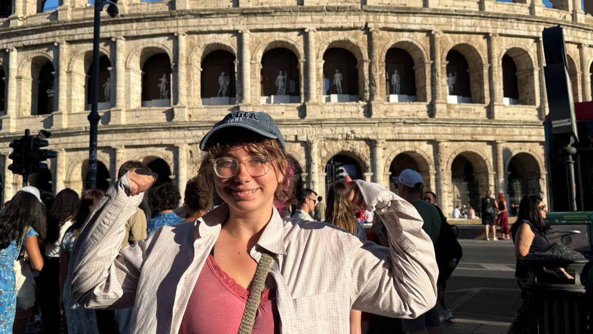 A young woman in a cap and glasses smiles at the camera with her arms raised in front of the ancient Roman Colosseum.