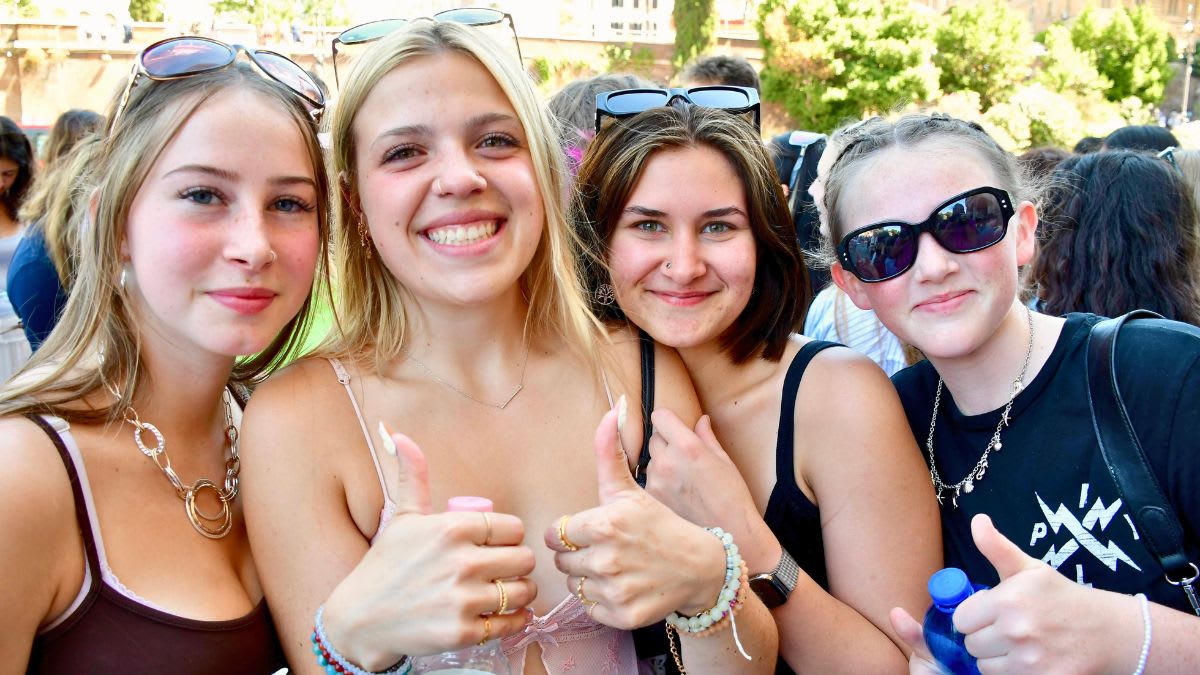 Four smiling young women with sunglasses pose for a photo in the foreground, with a blurred crowd and greenery in the background.