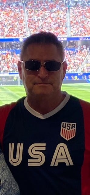 A man wearing a USA soccer jersey and sunglasses stands in the foreground with a blurred stadium crowd in the background.