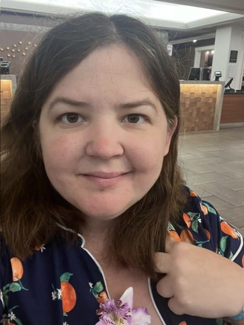 A woman with brown hair and a floral pajama top smiles at the camera in the foreground, with a blurred hotel lobby visible in the background.