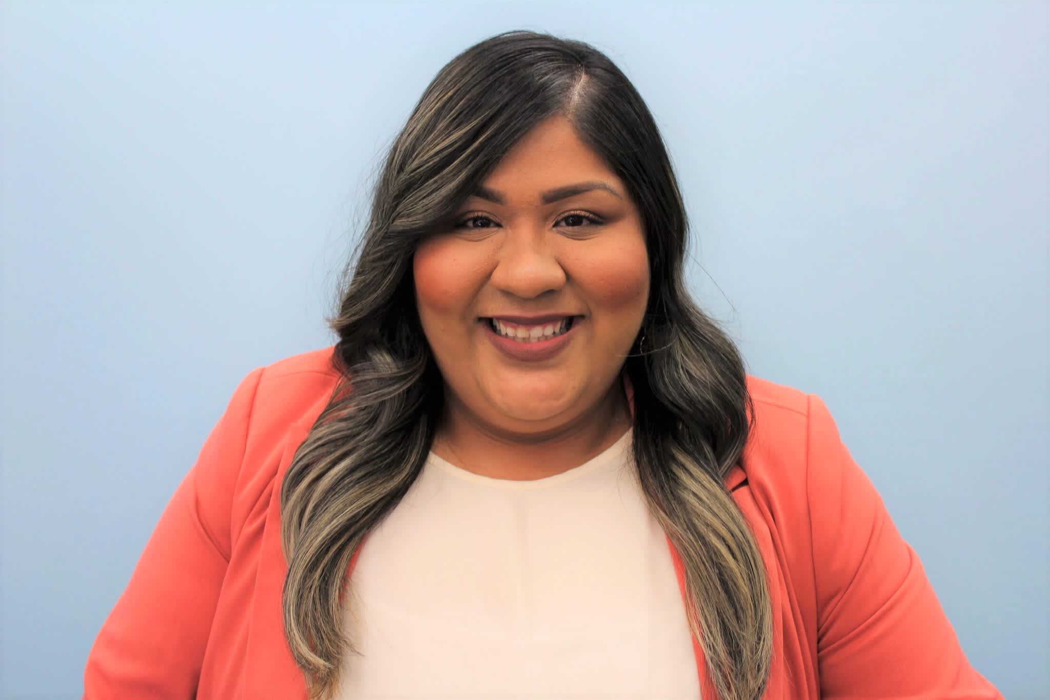 A smiling woman in a coral blazer and cream top is centered against a soft blue background.