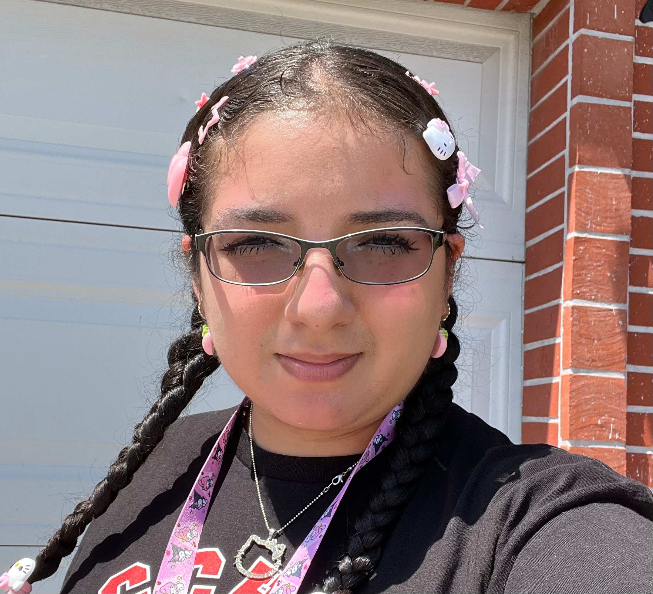 A young woman with braided hair adorned with pink bows and Hello Kitty clips smiles at the camera, with a white garage door and brick wall in the background.