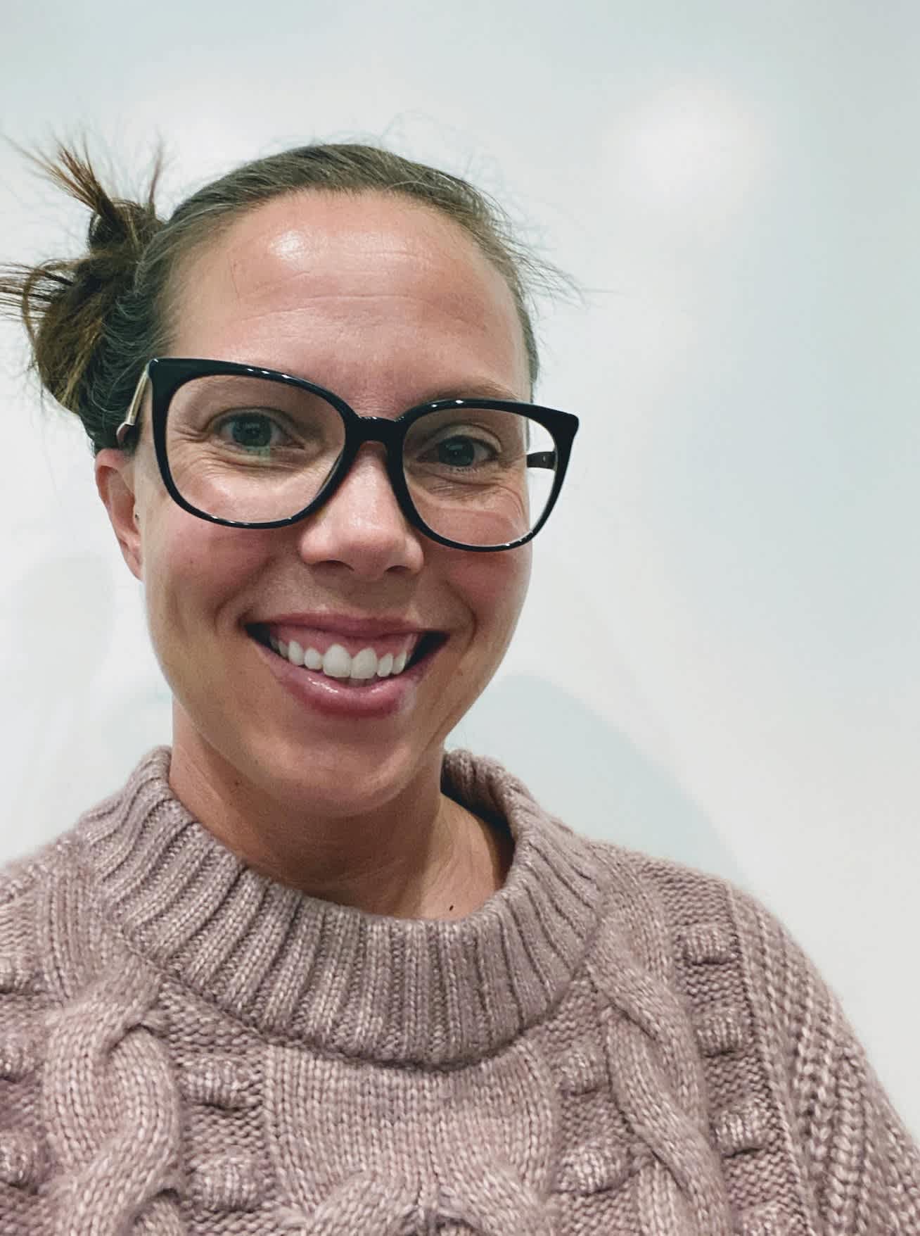 A woman with glasses and a cable-knit sweater smiles in the foreground against a plain white background.