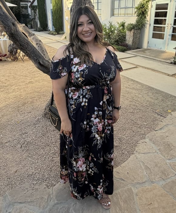 A woman in a floral dress stands on a gravel path in front of a building with a large tree to her left.