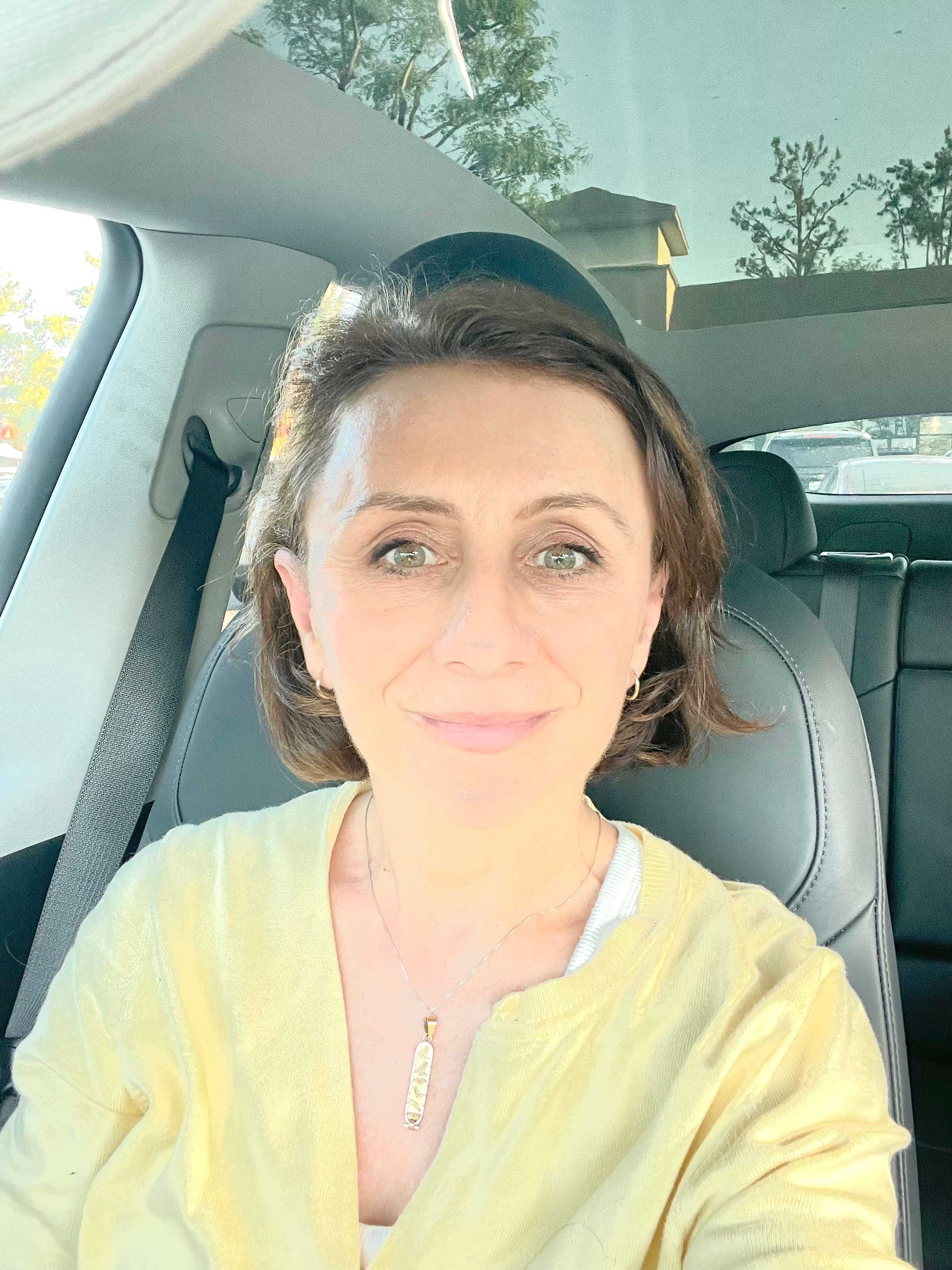 A woman with short brown hair and a yellow shirt smiles from the driver's seat of a car, with trees and buildings visible in the background.