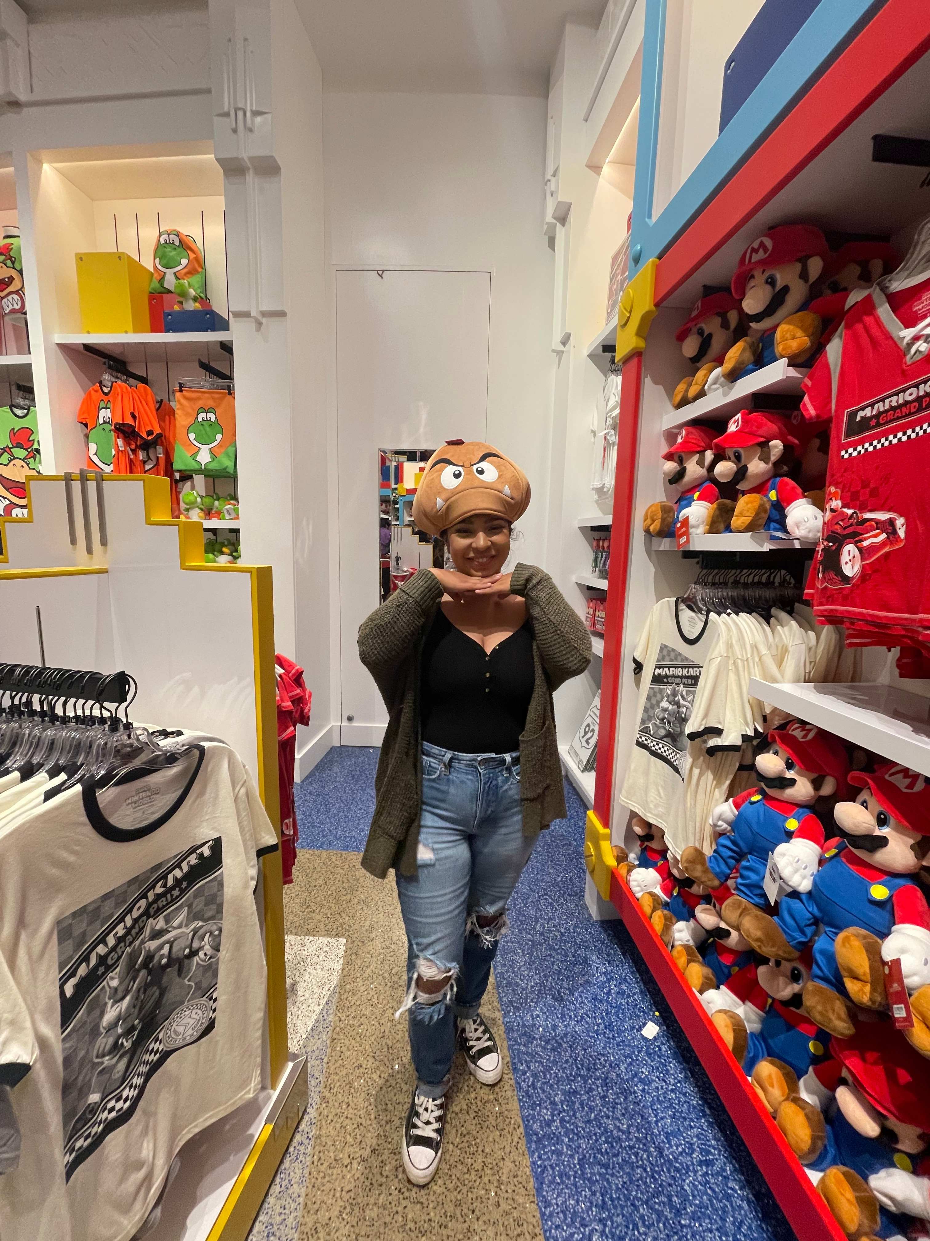 A person wearing a Goomba hat poses in a store aisle filled with Mario-themed merchandise.