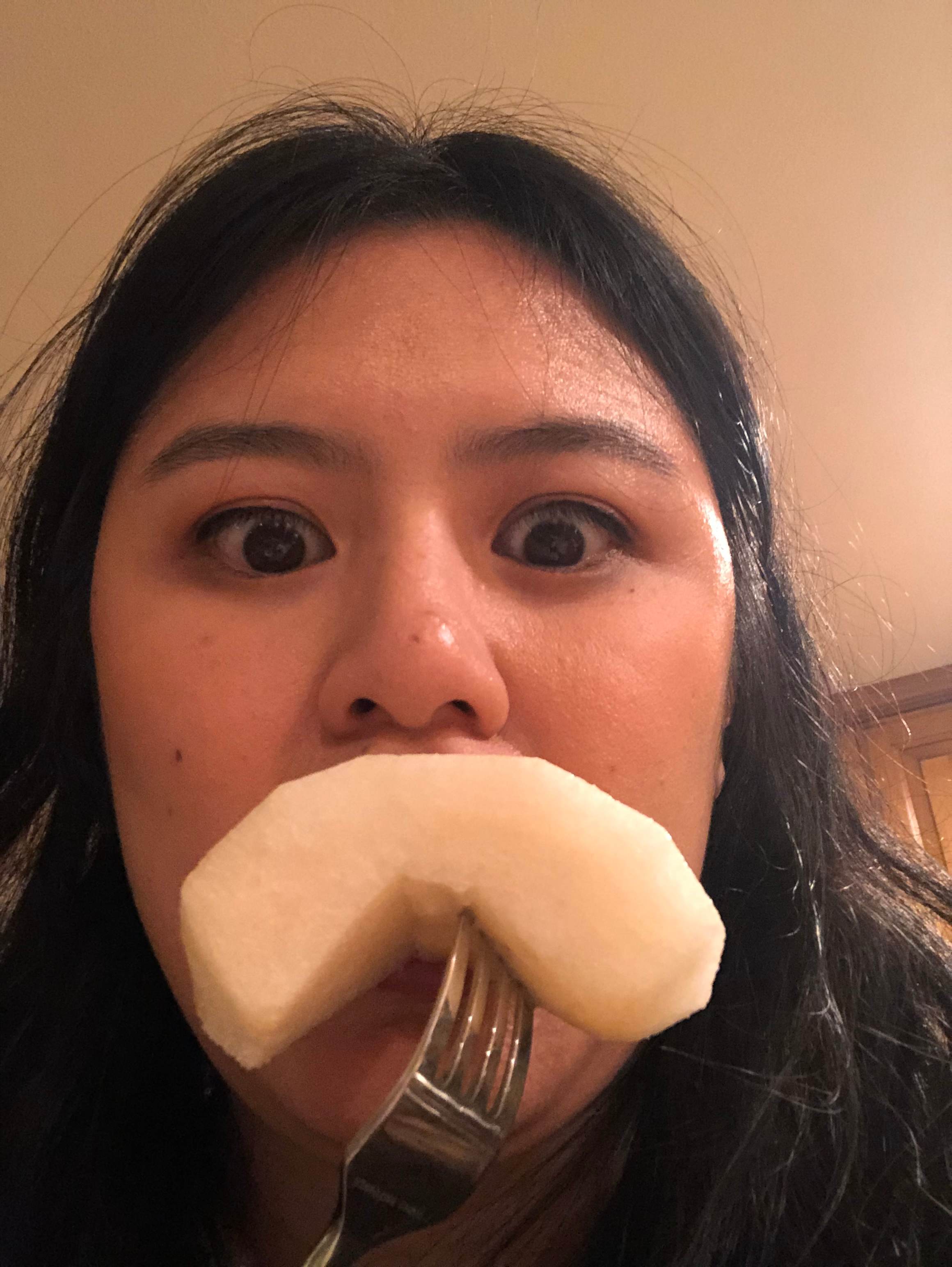 A young woman with wide eyes holds a slice of pear on a fork in front of her mouth, with a blurred kitchen background behind her.
