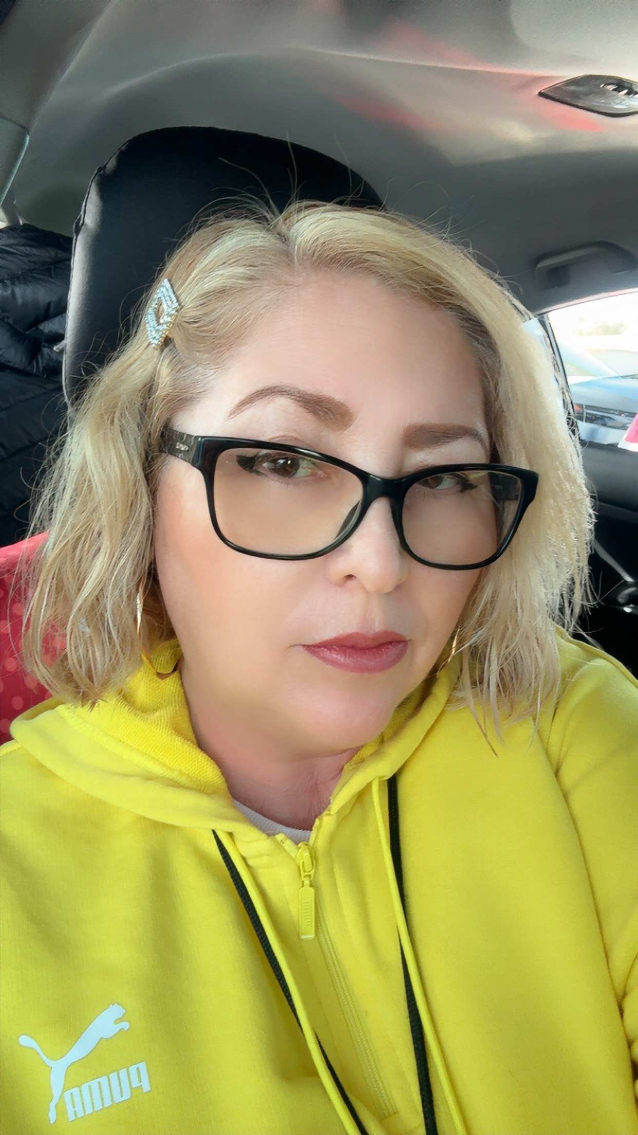A woman with blonde hair and glasses, wearing a yellow hoodie, is in the foreground, while the background shows the interior of a car.