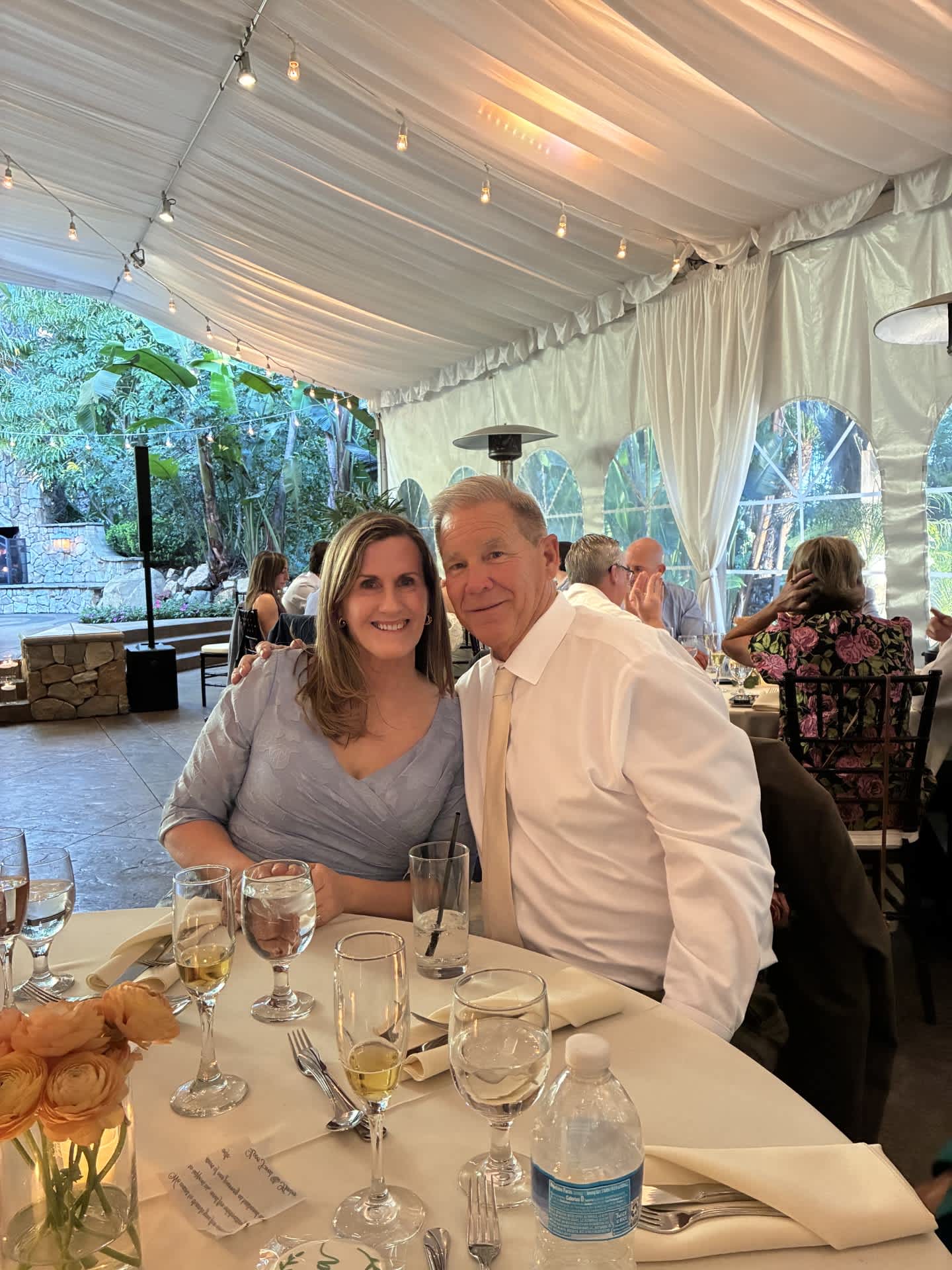A smiling couple poses at a formal dinner table set with elegant glassware and floral arrangements, while other guests are visible in the background under a white tent with lush greenery peeking through.