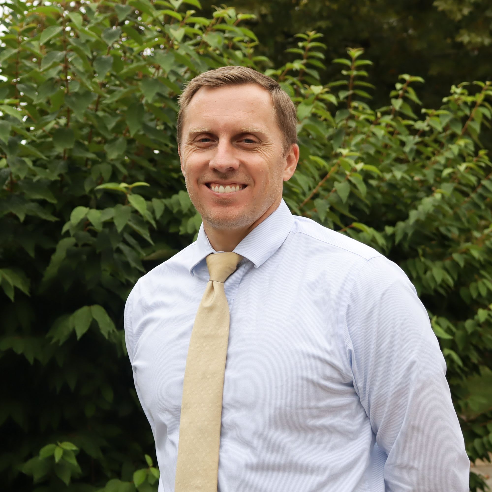 A smiling man in a light blue shirt and tan tie stands in front of a lush green bush.