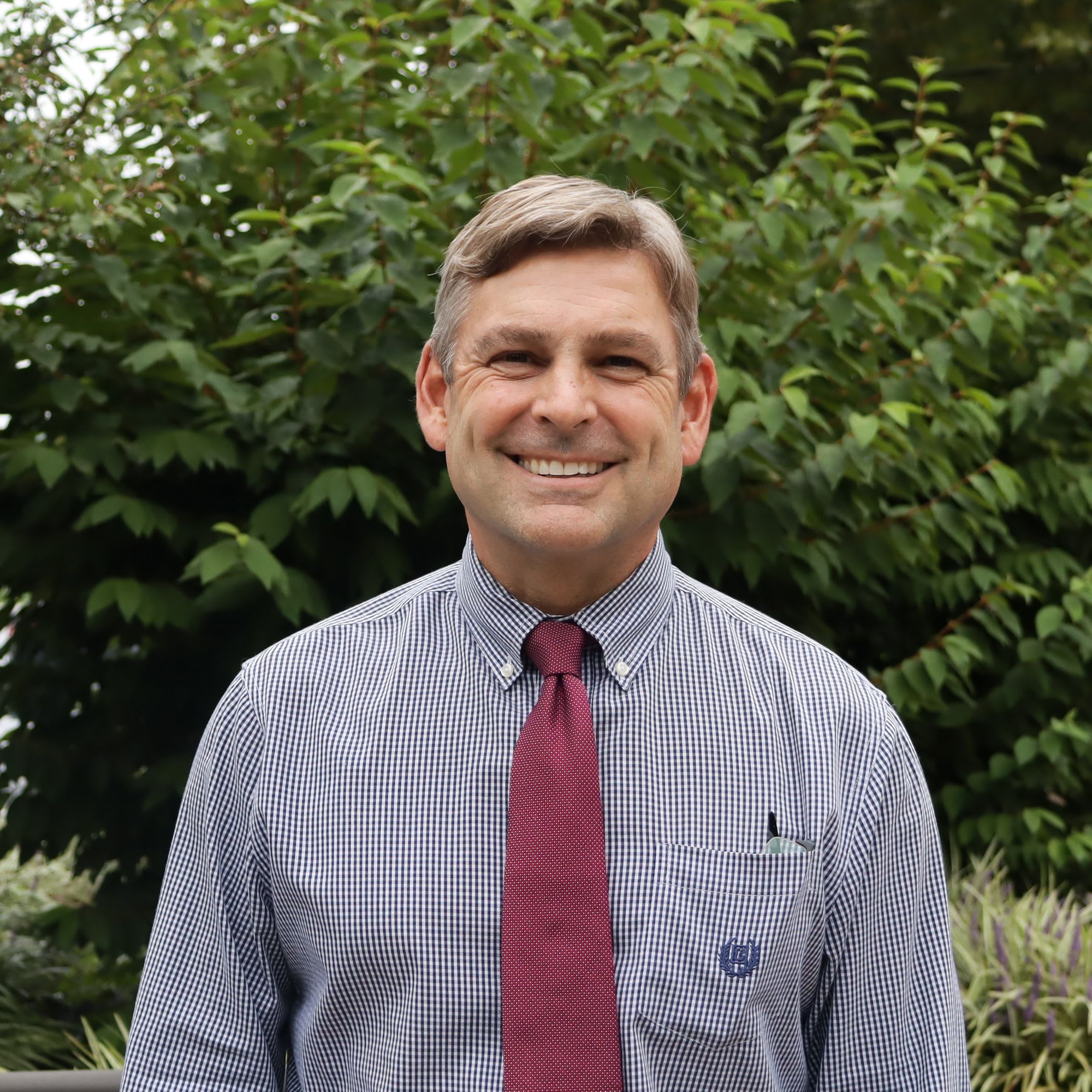 A smiling man in a collared shirt and tie stands in front of lush green foliage.
