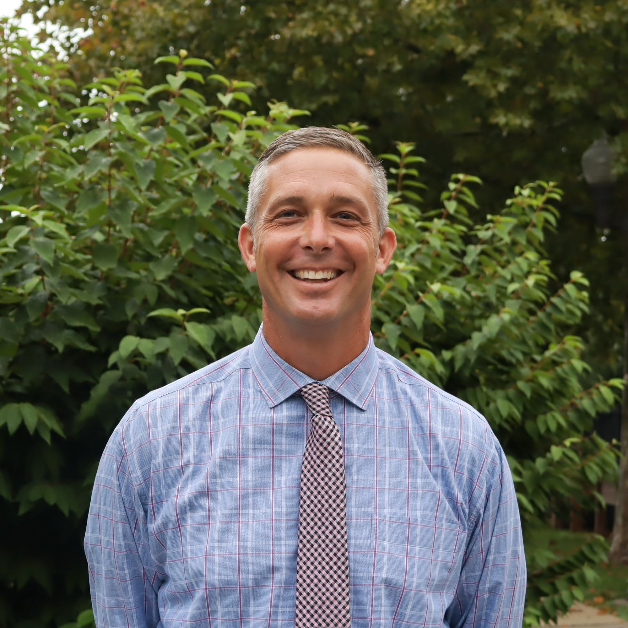 A smiling man in a blue plaid shirt and tie stands in front of lush green foliage.
