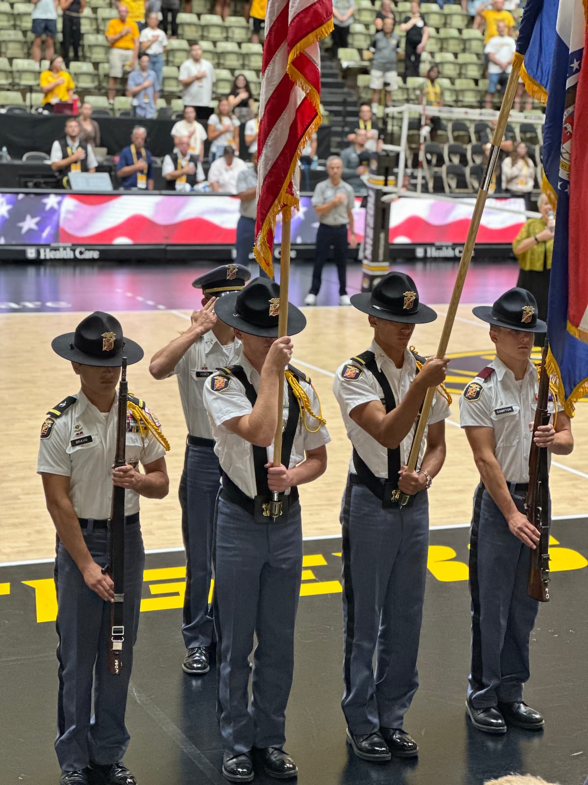 MMA cadets in uniform stand at attention while bearing flags in an indoor stadium