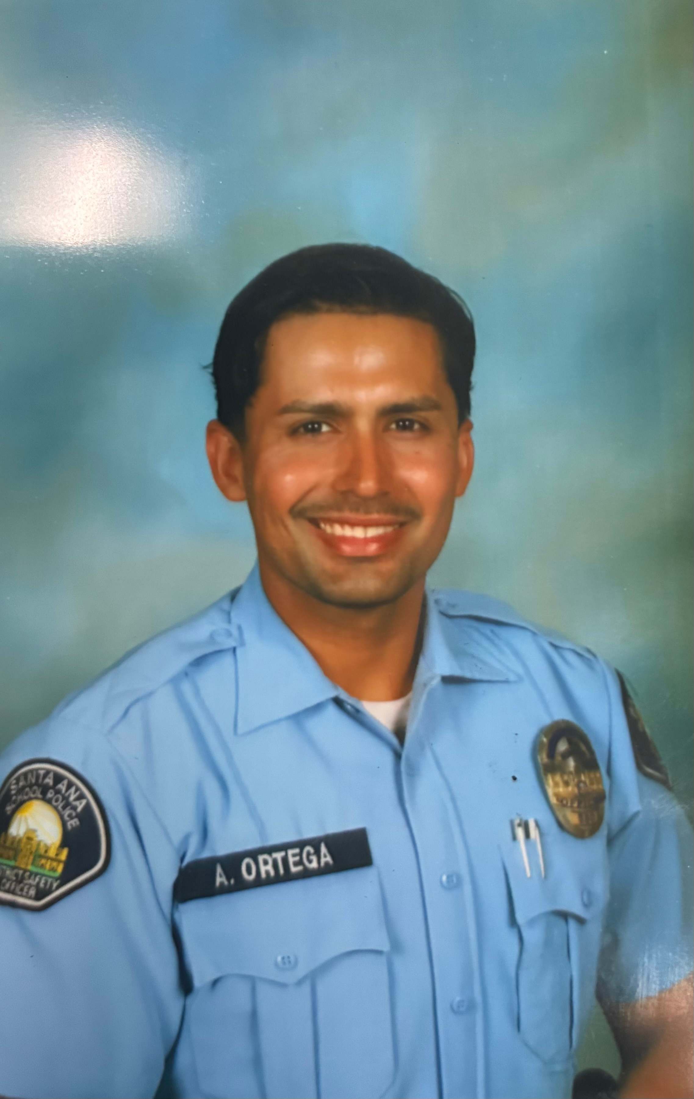 A smiling man in a light blue police uniform stands against a soft, blurred blue background.