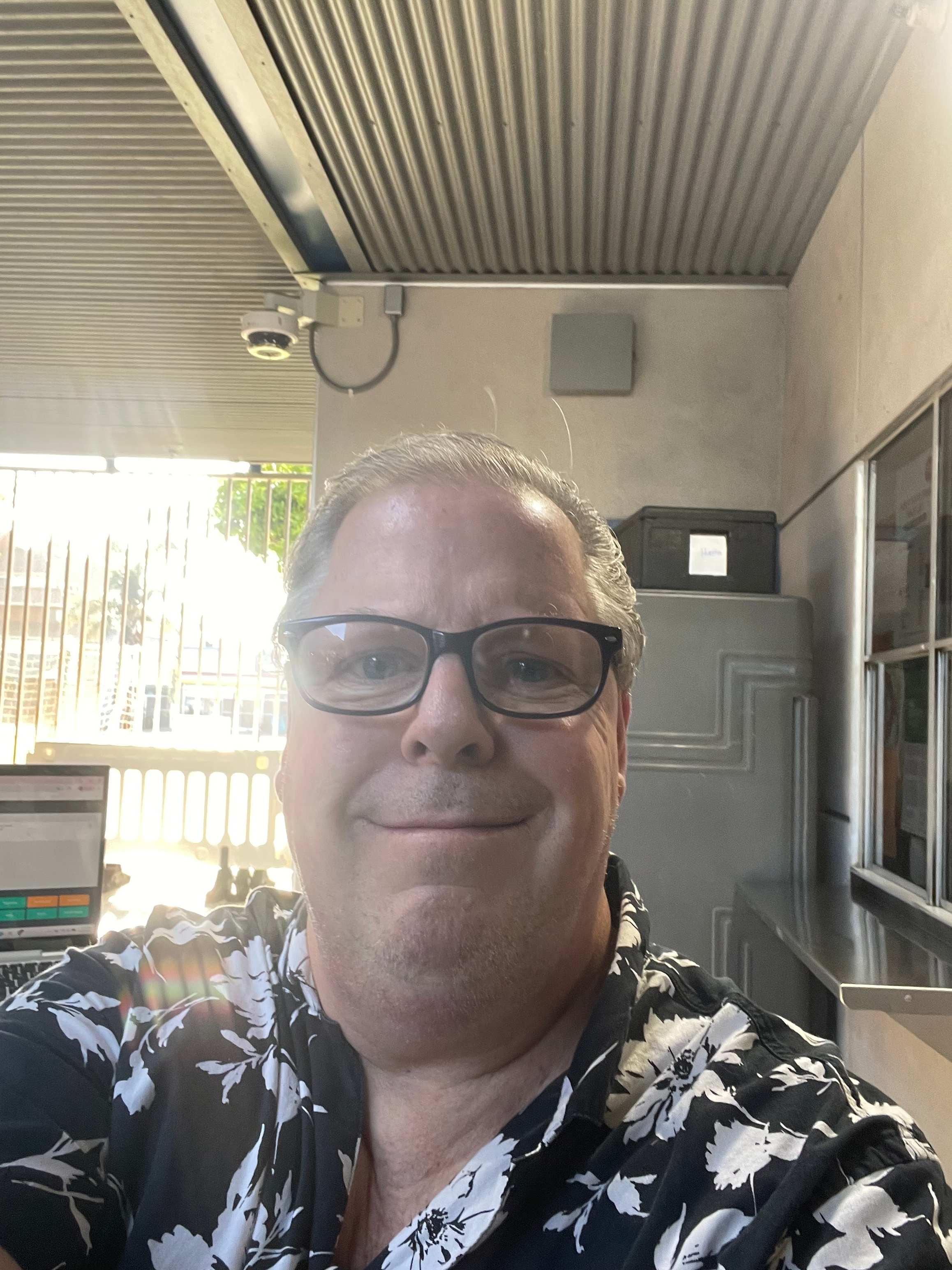 A man wearing glasses and a black and white floral shirt stands in the foreground against a background of a corrugated metal ceiling and a grey wall.