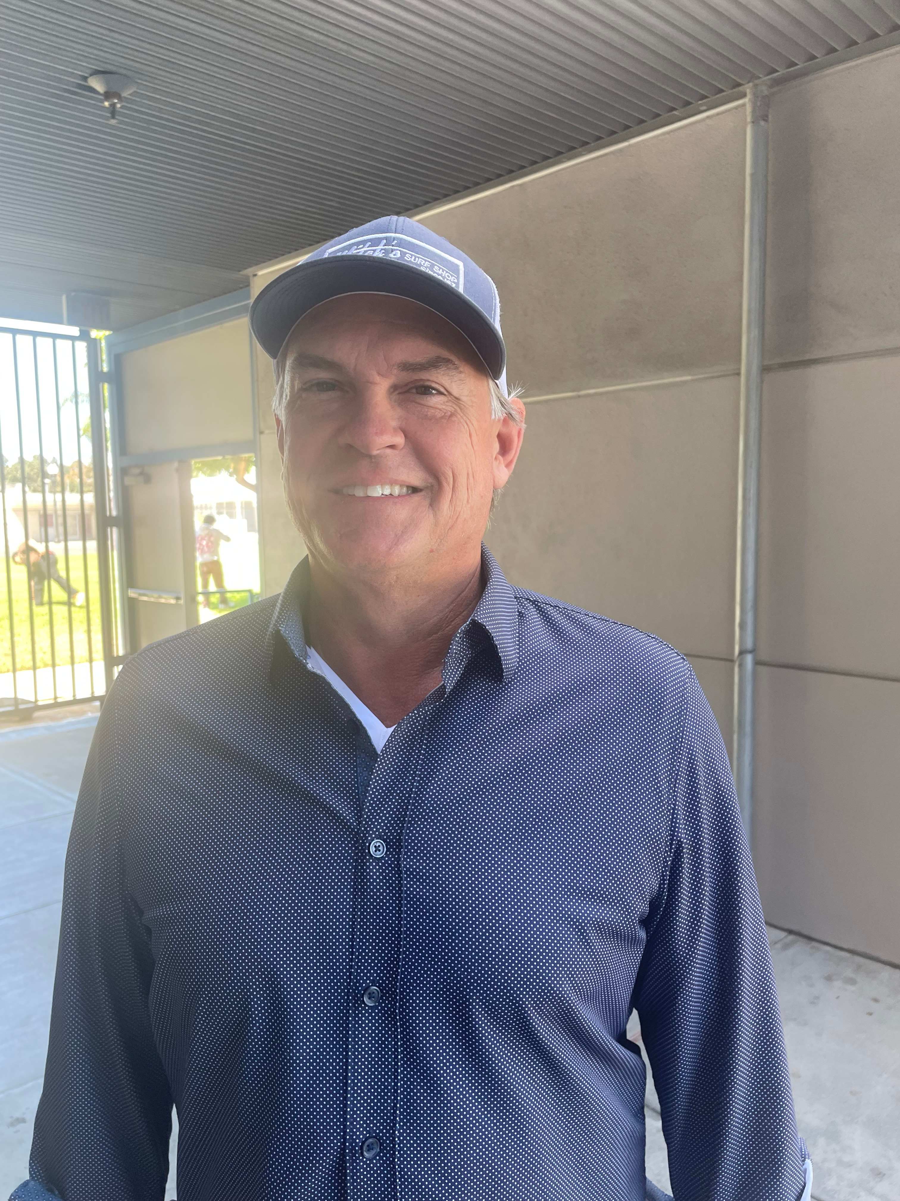 A smiling man wearing a blue button-down shirt and a baseball cap stands in the foreground against a background of a tan wall and a corrugated metal ceiling.