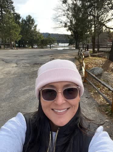 A woman wearing a pink beanie and sunglasses smiles in the foreground, with a paved path, trees, and a distant body of water in the background.