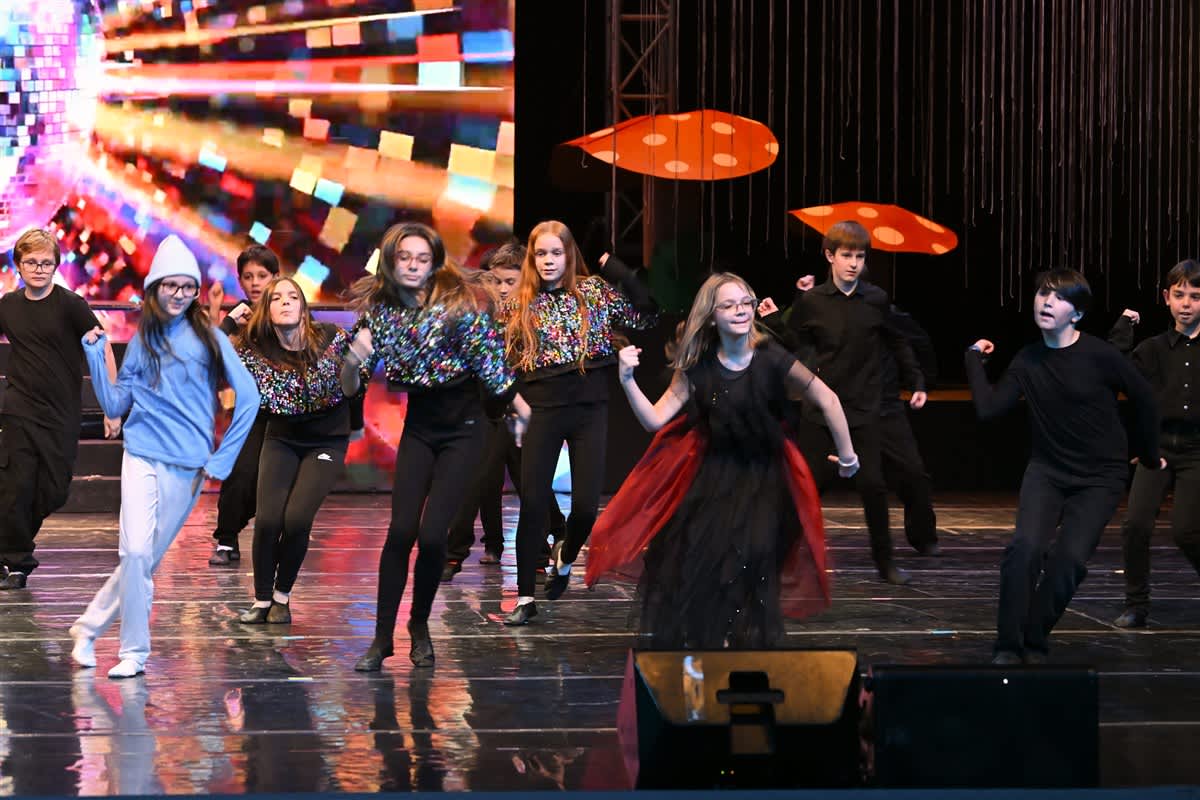 A group of children in various costumes dance on a reflective stage in front of a colorful digital screen and dark curtains.