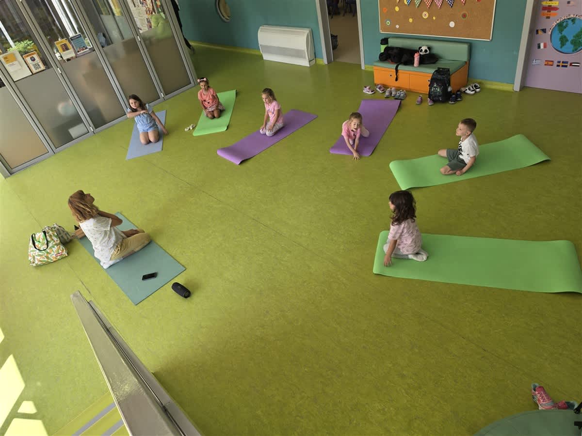 Children practice yoga on colorful mats in a bright room with a green floor and a storage bench in the background.