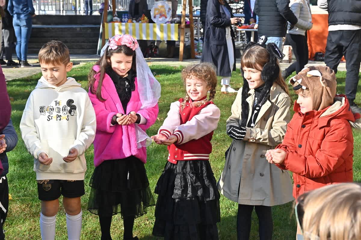 Five children in various costumes stand on a grassy lawn with people and a yellow-striped stand in the background.
