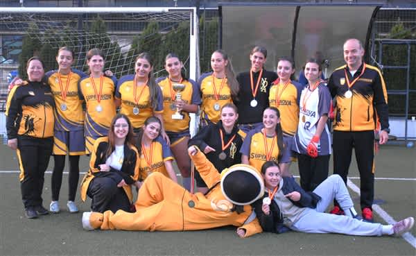A group of athletes in yellow and black uniforms pose with medals and a trophy on a sports field in front of a goal net.