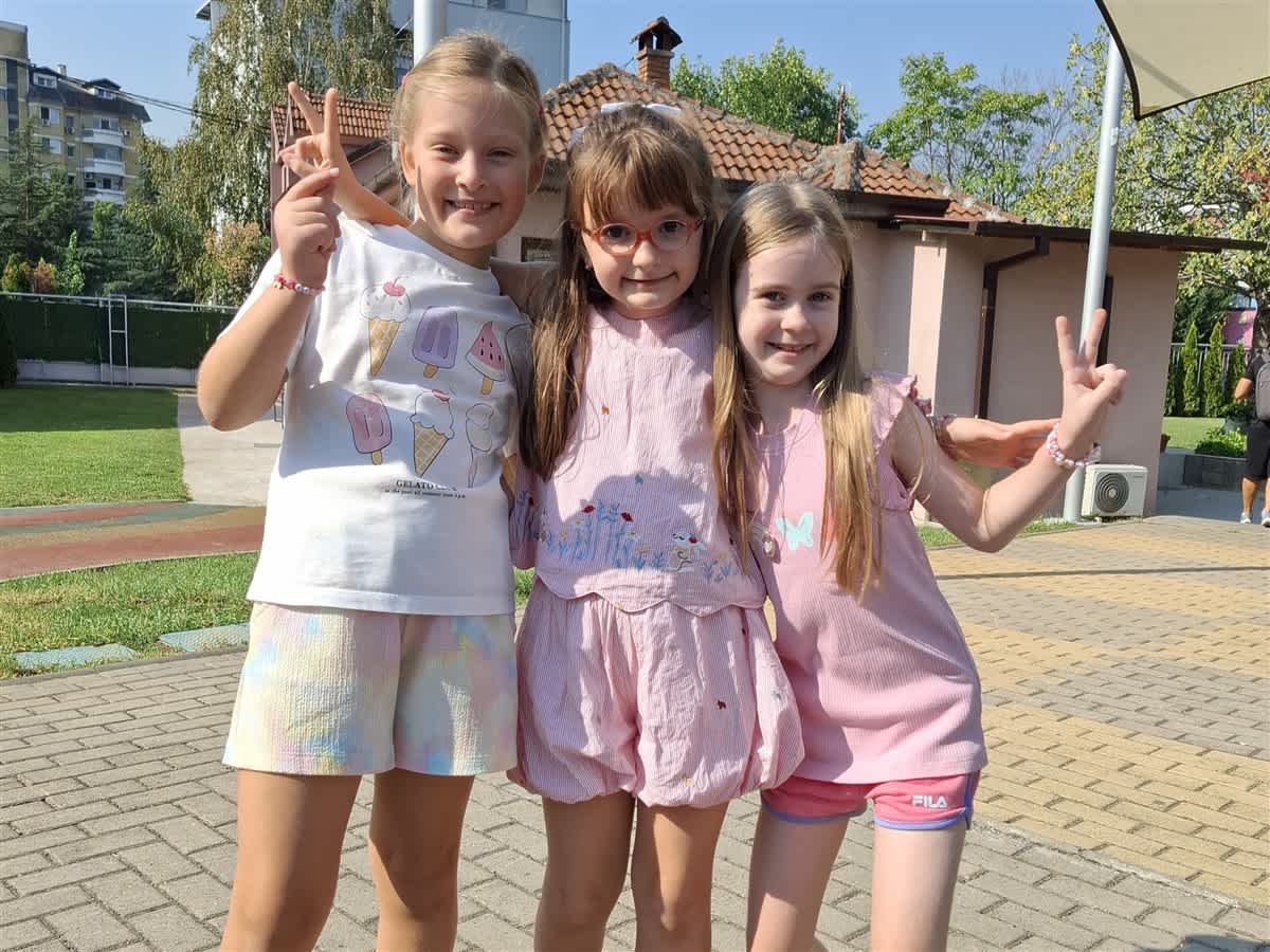 Three young girls smile and make peace signs in the foreground, standing on a paved patio with a house and green lawn in the background.