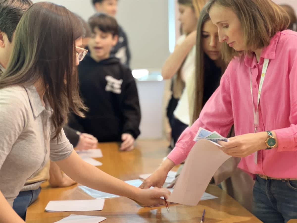 A woman in a pink shirt handles papers at a table while several students stand and watch in the background.