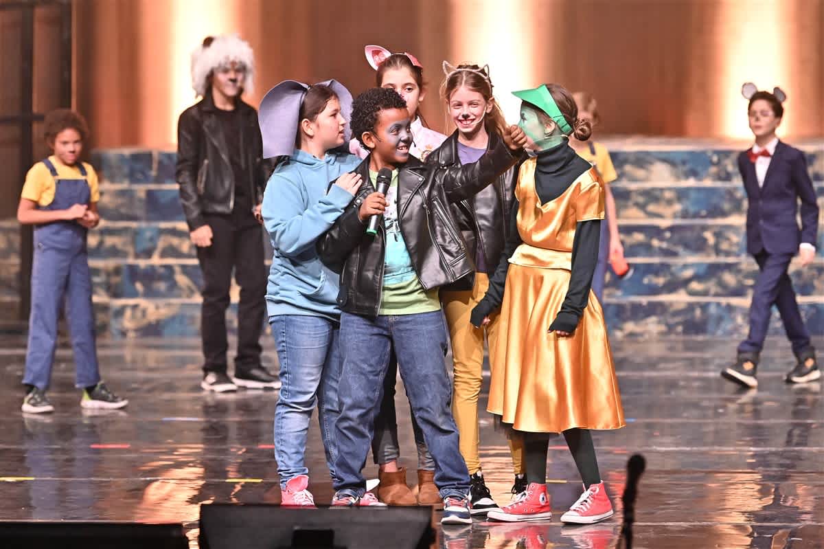 A group of children in various costumes perform on a glossy stage against a backdrop of textured blue and gold walls.