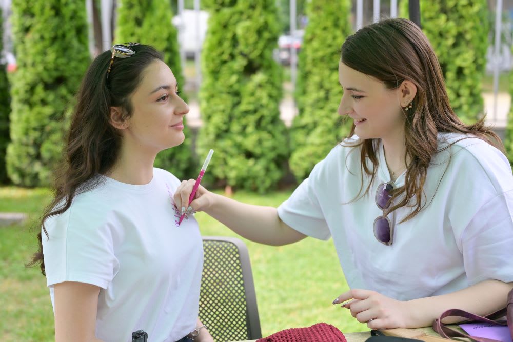 Two students smiling as one paints a design on the other's shirt outdoors