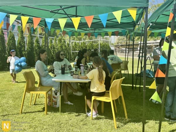 People sit at a white table under a green canopy decorated with colorful bunting, with a grassy yard and trees in the background.
