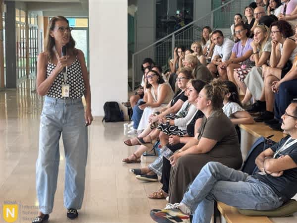 A woman stands speaking into a microphone in the foreground while a group of people sit and listen on stairs in the background.