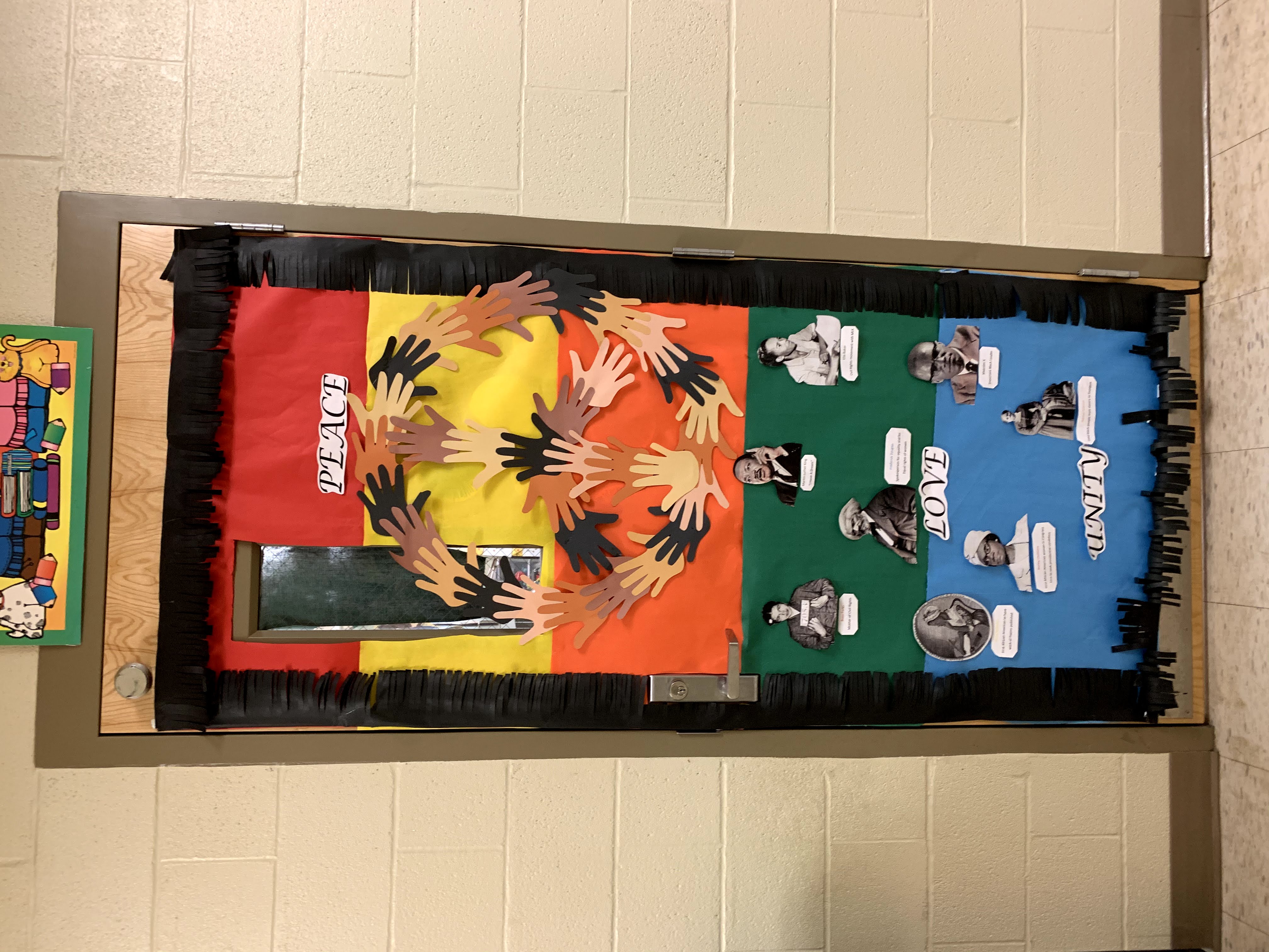 A classroom door is decorated with colorful paper, a peace sign made of hand cutouts, and images of historical figures, set against a brick wall.